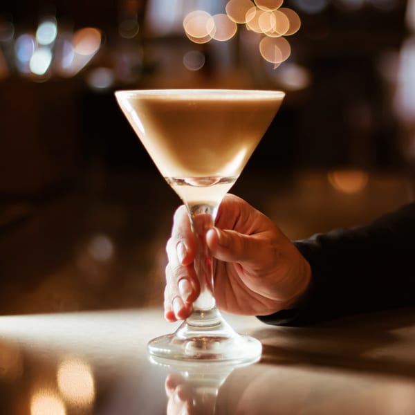 A hand holds a creamy cocktail in a martini glass at Flavio Restaurant DC, with bokeh lights in the background.