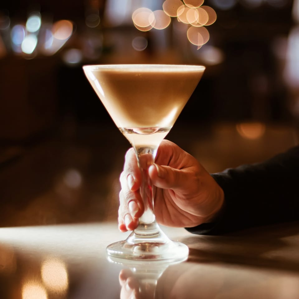 A hand holds a creamy cocktail in a martini glass at Flavio Restaurant DC, with bokeh lights in the background.