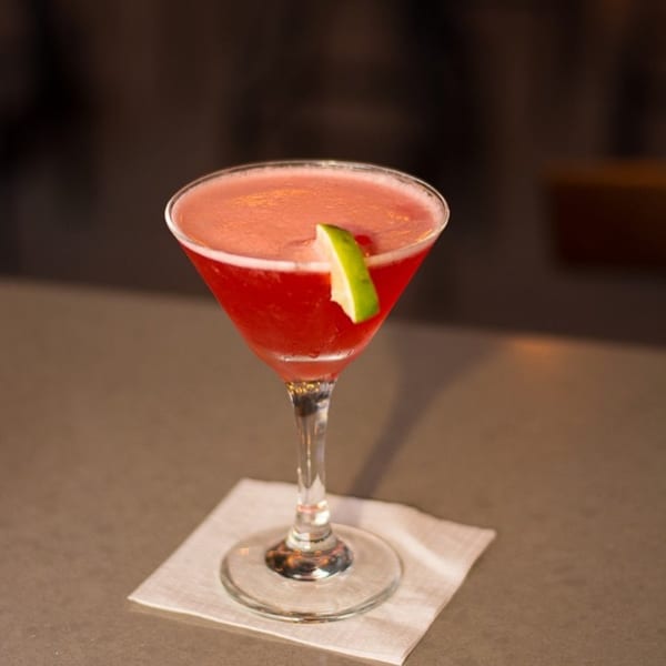 A vibrant red cocktail in a martini glass, garnished with a fresh lime wedge, on a bar.